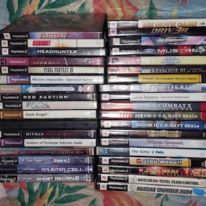 May include: A collection of PlayStation 2 video game cases, stacked in two columns. Titles include "Flatout", "Burnout", "Hitman Blood Money", "Star Wars", and "NASCAR Thunder 2004". The cases are in various colors.