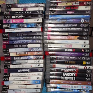 May include: A collection of PlayStation 3 video game cases arranged in two vertical stacks. The spines display various titles including Tomb Raider, Heavy Rain, Skyrim, Dragon Age, Battlefield, and multiple Assassin's Creed game editions.