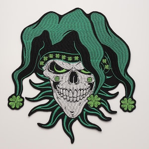 May include: An embroidered patch featuring a white skull wearing a green jester hat with four-leaf clover accents. The skull has green eye sockets and a wide grin, surrounded by green clover motifs and a black border on a white background.