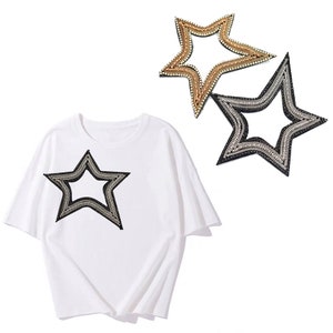 以下が含まれることがあります： 白いTシャツに、大きく黒と銀の星型のラインストーンの装飾が施されています。星型の装飾が2つ、別々に表示されています。1つは金と銀、もう1つは黒と銀です。