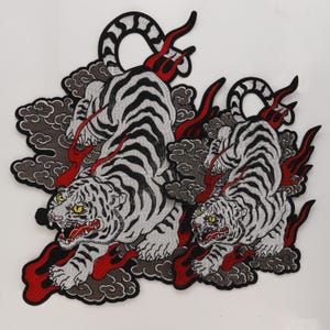 Puede incluir: Un parche bordado que muestra dos tigres blancos con rayas negras, ojos amarillos y bocas abiertas. Los tigres están rodeados de nubes grises y detalles rojos en forma de llamas. El diseño está delineado con hilo negro sobre fondo blanco.