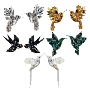 Puede incluir: Una colección de pendientes con forma de colibrí en varios estilos. Los pendientes presentan intrincados abalorios y detalles de cristal en plata, oro, negro, verde y blanco. Algunos diseños incluyen detalles de cadena colgante, mostrando una gama de texturas y colores.