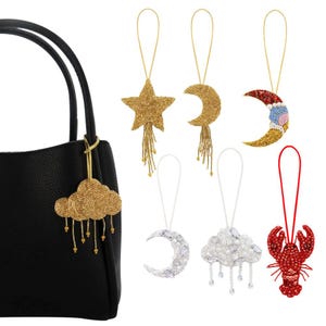 May include: A black handbag with a gold cloud charm. Other beaded charms include a gold star, crescent moon, and a red lobster. Additional charms are white and silver. All charms are beaded.