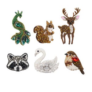Könnte beinhalten: Eine Sammlung von sechs Tierbroschen mit Perlen. Die Broschen umfassen einen grünen Pfau, ein braunes Eichhörnchen, einen braunen Hirsch mit rosa Akzenten, einen schwarz-weißen Waschbären, einen weißen Schwan und einen braunen Vogel mit einem roten Edelstein.