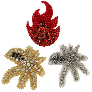 May include: Three rhinestone embellishments in the shapes of a flame, a starburst, and a palm tree. The flame is red, the starburst is silver, and the palm tree is gold.