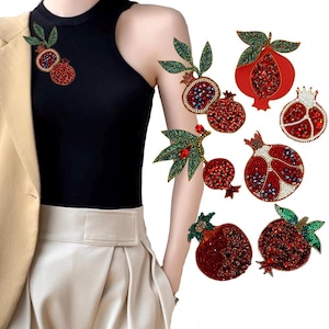 Puede incluir: Un conjunto de siete broches de granada rojos y verdes con joyas. Los broches están hechos de pedrería y cuentas y presentan un diseño detallado.