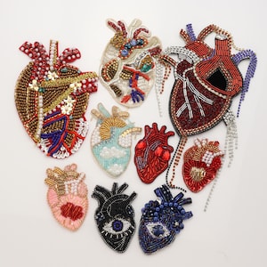 Può includere: Una collezione di nove toppe a forma di cuore anatomico realizzate con intricate lavorazioni di perline, paillettes e strass. I design presentano varie palette di colori, tra cui rosso, blu, nero, oro e rosa pastello, alcuni con motivi a occhio.