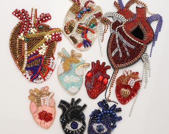 Puede incluir: Una colección de nueve parches de corazones anatómicos elaborados con intrincados trabajos de cuentas, lentejuelas y pedrería. Los diseños presentan varias paletas de colores, incluyendo rojo, azul, negro, dorado y rosa pastel, con motivos oculares.