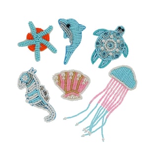 Può includere: Un set di sei applicazioni di creature marine in perline. Le applicazioni sono realizzate con perline blu, rosa e bianche e presentano una stella marina, un delfino, una tartaruga, un cavalluccio marino, una conchiglia e una medusa.