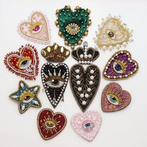 May include: A collection of twelve decorative embroidered patches featuring heart and star shapes. The designs incorporate intricate beadwork, sequins, pearls, and metallic threads, with several pieces displaying detailed eye motifs and crown accents.