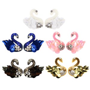 May include: An assortment of decorative swan embellishments. Each pair features a swan design with sequins and rhinestones. Colors include white, blue, pink, black, and gold. These embellishments are suitable for various craft projects.