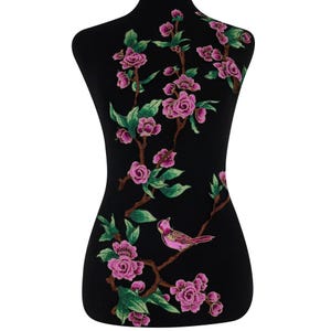 Op de afbeelding: Zwarte paspop met een bloemenontwerp. Het ontwerp toont roze bloemen, groene bladeren en een roze vogel die op een bruine tak zit. De bloemen zijn geborduurd, wat een textuur geeft.