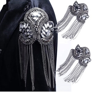 Puede incluir: Dos adornos de cadena plateada y pedrería con cadenas colgantes. Los adornos tienen la forma de una flor estilizada con una piedra de pedrería central.
