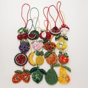 Può includere: Una collezione di ornamenti a forma di frutta e verdura realizzati con perline e paillettes colorate. Ogni pezzo è dotato di un occhiello per appenderlo e raffigura elementi tra cui ciliegie, fette di anguria, un fungo, uva, una pesca, zucca, pera, banana, ananas, arancia e lime.