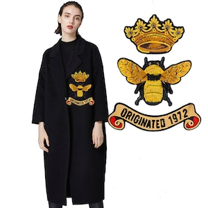 Op de afbeelding: Drie geborduurde strijkpatches met een bij, een kroon en een banner met de tekst "Originated 1972". De patches zijn geel en goudkleurig met zwarte contouren.