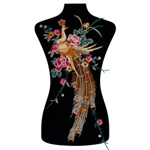 Puede incluir: Parche de pavo real bordado con un diseño detallado. El pavo real es en tonos dorados, naranjas y marrones, rodeado de flores rosas y naranjas y hojas verdes. El parche está sobre un fondo negro.