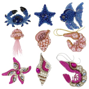 May include: Assortment of colorful beaded iron-on patches featuring a variety of sea life and nautical themes. The patches include a blue crab, a blue starfish, a blue fish, a pink seashell, a pink jellyfish, a pink lobster, a pink seashell, and a pink lobster.
