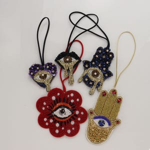 May include: Five decorative ornaments featuring intricate beadwork. Designs include a purple heart with an eye, a black lip shape with an eye, a dark blue hamsa with an eye, a red flower with a central eye, and a gold hamsa hand with a blue eye.