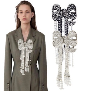 Può includere: Un paio di grandi spille decorative a forma di fiocco decorate con gioielli. Una spilla è nera con strass trasparenti, l'altra è bianca con strass trasparenti. Entrambe le spille hanno lunghe catene di perline pendenti.