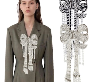 Puede incluir: Un par de grandes broches decorativas con forma de lazo adornadas con joyas. Una broche es negra con diamantes de imitación transparentes, la otra es blanca con diamantes de imitación transparentes. Ambas broches tienen largas cadenas de cuentas colgantes.