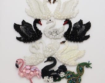 Puede incluir: Una colección de parches bordados con diversas aves, incluyendo cisnes blancos y negros, un flamenco rosa y un pavo real verde. Cada parche está adornado con cuentas, lentejuelas y cristales, mostrando texturas intrincadas y diseños detallados.