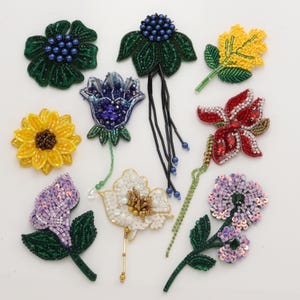 May include: A collection of eight handcrafted floral brooches featuring intricate beadwork. The set includes various flower shapes in vibrant colours such as red, yellow, white, purple, and green, with some designs incorporating long beaded fringe accents.