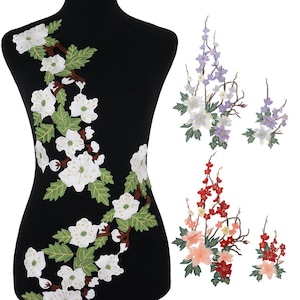 Puede incluir: Apliques florales bordados en blanco, morado y rojo. El aplique más grande es un diseño de flores blancas sobre un maniquí negro. Los apliques adicionales incluyen racimos de flores moradas y rojas con hojas verdes.