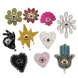 Puede incluir: Colección de broches decorativos en varias formas, incluyendo flores, una concha, una estrella, un corazón, una mano y una mano de Fátima. Cada broche presenta un diseño de ojo con intrincados abalorios y acentos coloridos. Los broches son de varios colores, incluyendo oro, plata, rosa y negro.