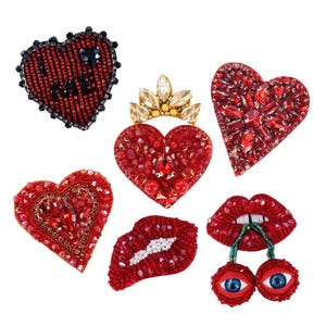 Könnte beinhalten: Eine Sammlung roter, mit Perlen besetzter Aufnäher in verschiedenen Formen, darunter Herzen, Lippen und ein Herz mit Krone. Die Aufnäher sind mit Perlen und Kristallen verziert, was einen funkelnden Effekt erzeugt. Ein Aufnäher hat das Wort "LOVE".