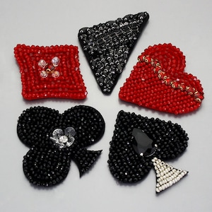 Könnte beinhalten: Fünf gestickte Filzpatches in Form von Spielkarten-Symbolen. Die Symbole sind Pik, Herz, Karo und zwei Kreuz. Die Patches sind schwarz und rot mit Strasssteinen.