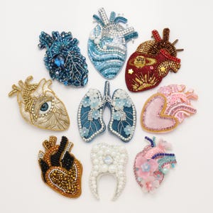 Puede incluir: Una colección de nueve broches con forma de órganos anatómicos, que incluye siete corazones, un par de pulmones y un diente. Cada pieza está intrincadamente decorada con diversas cuentas, lentejuelas, cristales y detalles florales de colores.