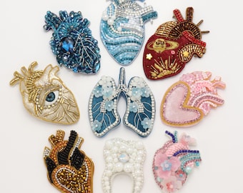 Puede incluir: Una colección de nueve broches con forma de órganos anatómicos, que incluye siete corazones, un par de pulmones y un diente. Cada pieza está intrincadamente decorada con diversas cuentas, lentejuelas, cristales y detalles florales de colores.