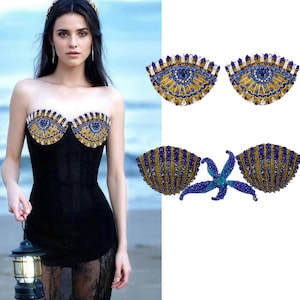 Könnte beinhalten: Ein schwarzes trägerloses Kleid mit einem dekorativen Oberteil mit einem Augendesign in Gold, Blau und Weiß. Das Bild zeigt auch zwei separate augenförmige Verzierungen und einen muschelförmigen BH mit einem Seestern.