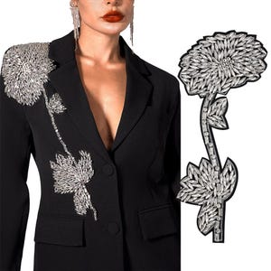 May include: A black blazer featuring a large, silver, rhinestone flower design on the left shoulder. The flower is crafted from numerous small, sparkling rhinestones.