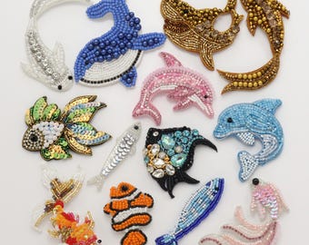 Puede incluir: Una colección de doce parches bordados con formas de diversas criaturas marinas, incluyendo ballenas, delfines y peces. Las piezas están elaboradas con cuentas, lentejuelas y cristales de colores, presentando tonos blancos, azules, dorados, rosas, naranjas y negros.
