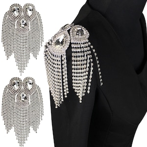 Puede incluir: Charreteras de hombro de diamantes de imitación plateados con una gran piedra central en forma de lágrima y flecos en cascada. Los adornos decorativos se muestran en una prenda negra, con dos charreteras adicionales mostradas por separado.
