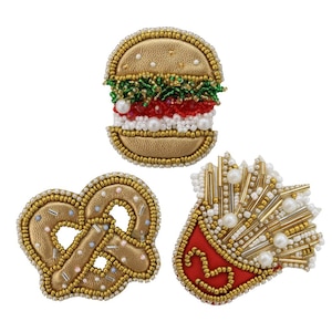 Może przedstawiać: Trzy haftowane naszywki o tematyce jedzenia: hamburger, precel i porcja frytek. Hamburger ma złoty bułkę, zieloną sałatę, czerwony pomidor i biały ser. Precel jest złoty z różowymi i niebieskimi posypkami. Frytki są złote z białymi perłami i czerwonym ketchupem.