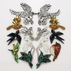 May include: A collection of bird-shaped patches featuring intricate beadwork, sequins, and crystals. The assortment includes silver, gold, black, white, and green designs, showcasing various avian forms with detailed textures and reflective embellishments.