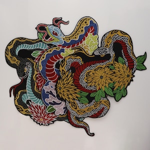 May include: A large, intricate embroidered patch featuring multiple intertwined snakes in vibrant colours including red, blue, yellow, and green. The design incorporates detailed floral elements, specifically large golden chrysanthemums and red blossoms, set against a black background with a thick black border.