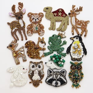 Peut inclure: Un ensemble de douze écussons en forme d'animaux ornés de perles et de paillettes. La collection comprend un cerf, un ours en peluche, un chameau, un écureuil, une grenouille couronnée, un pingouin, un lapin, un hibou, un hérisson et un raton laveur.