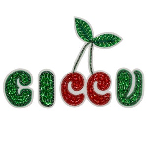 Può includere: Una toppa ricamata verde e rossa con la parola "cherry" scritta in paillettes. Le ciliegie sono rosse e le foglie sono verdi.