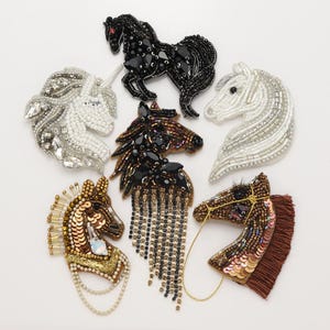 Könnte beinhalten: Eine Sammlung von sechs pferde- und einhornförmigen Broschen, die mit aufwendigen Perlenarbeiten, Pailletten und Kristallen verziert sind. Die Designs zeigen verschiedene Farbpaletten in Schwarz, Weiß, Gold und Braun, teilweise mit Fransen.
