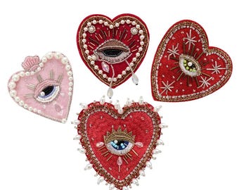 May include: Four embroidered felt heart-shaped patches with beaded details. Each patch features a large eye with a crown and radiating lines. The eyes are blue, green, and brown. The hearts are red and pink.