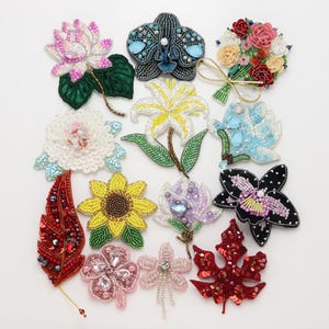 May include: A collection of thirteen handcrafted beaded brooches featuring various floral and botanical designs. The assortment includes depictions of a lotus, orchids, a sunflower, a bouquet, leaves, and clover shapes in vibrant colours.