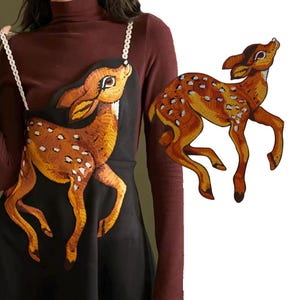 May include: A black dress with a brown and white embroidered deer design. The dress has a scoop neck and a fitted bodice. The deer is facing to the right and has a long neck and tail.