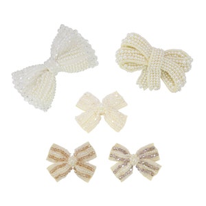May include: Five decorative bows in various designs. The bows are embellished with small, round, off-white beads. Two bows are a light cream colour, and two are a light tan colour. The fifth bow is a light cream colour with clear beads.