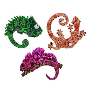 May include: Three beaded reptile brooches. One green chameleon with sequins, one orange gecko with floral accents, and one pink chameleon with gemstone embellishments. Each brooch is intricately detailed and set against a white background.