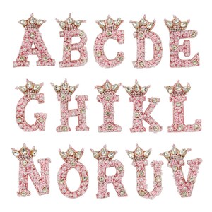 Może przedstawiać: Różowe filcowe litery alfabetu od A do V, każda z koroną z kryształków i pereł na górze.