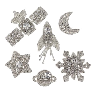 May include: A set of seven silver-toned, rhinestone-encrusted iron-on patches featuring space-themed designs, including a rocket, a satellite, a star, a crescent moon, a snowflake, and a planet.