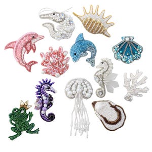 Op de afbeelding: Een verzameling van met kralen versierde versieringen met oceaanthema, waaronder een roze dolfijn, blauwe dolfijn, zeepaardje, garnaal, kwal en koraal. De versieringen zijn in verschillende kleuren, waaronder roze, blauw, wit en goud. Een groene kikker met een kroon is ook inbegrepen.